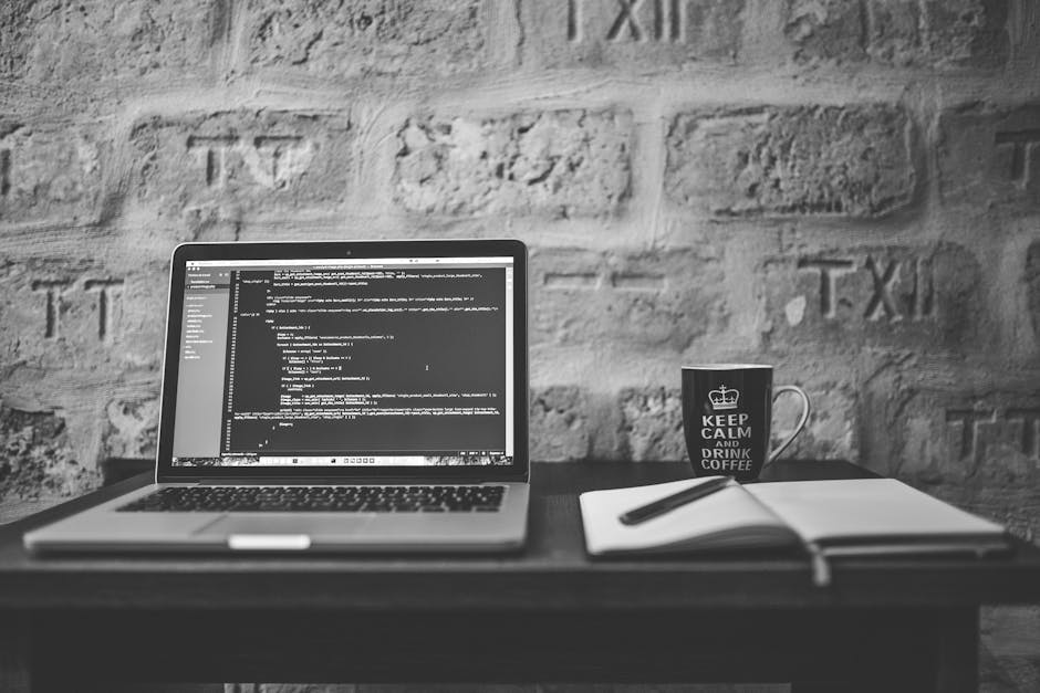 A black and white photo showing a workspace with a laptop displaying lines of code, positioned on a wooden desk against a textured brick wall. To the right of the laptop, there is a white notepad with