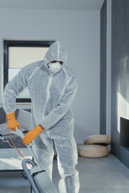 Close-up image of a black vacuum cleaner head actively cleaning a patterned area rug on a wooden floor in a well-lit room, with a person wearing grey pants and white shoes standing nearby. The rug fea